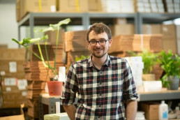 Man with glasses and plaid shirt standing in a warehouse with plant and boxes